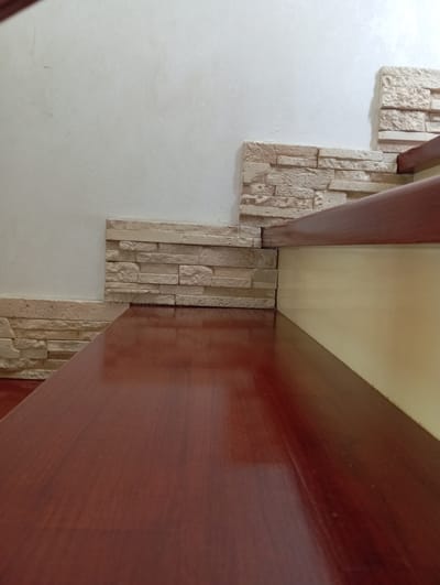 The image displays a close-up view of a section of a staircase. The foreground is dominated by a polished, dark red wooden surface, likely a stair tread, which reflects light and shows subtle wood grain. To the left and behind this surface, a wall is visible, decorated with stacked stone veneer tiles in shades of beige and cream, creating a textured accent. The tiles are arranged horizontally in staggered rows. The upper right portion of the image shows another dark red wooden element, possibly the handrail or a riser, positioned at an angle. A plain, textured off-white wall forms the backdrop in the upper left quadrant. The lighting appears to be ambient, casting soft shadows and highlighting the reflective surfaces. The overall scene suggests an interior architectural detail, possibly in a residential or commercial setting. The image does not provide any contextual information or visual cues to determine its location in Hlynytsi, Ukraine.