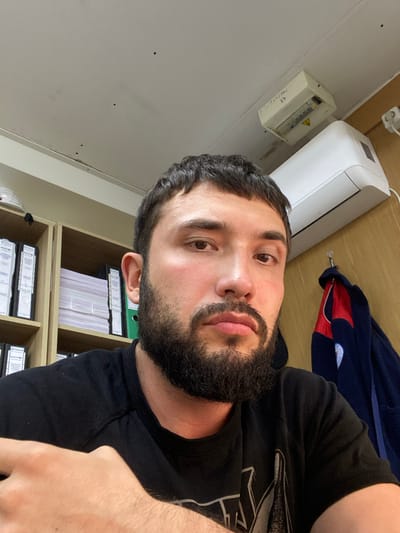 The image is a selfie taken by a man with a beard and short dark hair. He is wearing a black t-shirt and is sitting in an office environment. Behind him, there are shelves filled with binders and folders. An air conditioning unit is mounted on the wall above him. A jacket is hanging on a hook to the right of the frame. The ceiling shows some small dark spots. There is a white electrical box mounted on the ceiling, with some writing on it. The overall lighting suggests an indoor setting. The location is identified as Luzhitsy, Russia.