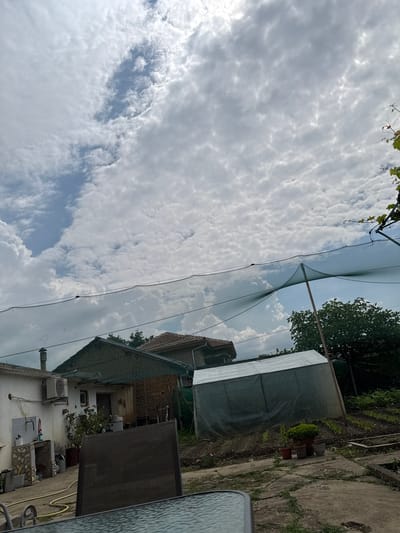 This image captures an outdoor scene in Hadzhidimitrovo, Bulgaria, depicting a residential backyard and garden area under a partially cloudy sky during daylight hours.
The upper half of the frame is dominated by the sky, exhibiting a mix of dense, white, stratocumulus clouds and interspersed patches of clear blue sky, particularly prominent on the left side and in a diagonal band through the upper middle.
In the foreground, the lower edge of a dark, reflective glass table is visible, with the top of a dark, mesh-backed chair occupying the lower-left quadrant. The ground consists of irregular concrete paving with visible cracks and sparse green weeds growing through them.
Moving into the midground from left to right:
A white, single-story building is positioned on the left, featuring a chimney on its roof and an external air conditioning unit mounted on its wall. Below the AC unit, an outdoor utility area includes a sink or wash basin with a faucet, surrounded by various buckets and pots. A coiled yellow hose lies on the concrete path nearby. Adjacent to this white building, a structure with a dark, possibly tiled, roof and textured stone or brick walls is visible. Further to the right, a large, rectangular structure resembling a greenhouse or storage shed is covered in a light-colored, translucent tarp or plastic material.
To the right of this structure, a cultivated garden plot is evident, with rows of small green plants growing in tilled earth. Several potted plants are placed on the concrete leading towards the garden. A large green tree is visible behind the greenhouse, and a dark green netting structure, supported by at least one wooden pole on the right, stretches horizontally across the upper midground, partially obscuring the sky and distant elements. This netting appears to be protective, possibly for crops or as a shade net. Green foliage, likely grapevines, is visible along the upper right edge, draped over a support.
No people are present in the scene, and no specific actions are occurring. The environment suggests a functional rural or semi-rural setting focused on domestic and gardening activities.