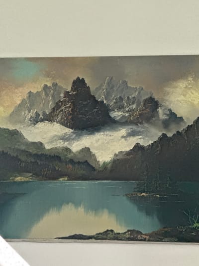 This is a painting of a serene mountain landscape with a calm lake in the foreground. The mountains are jagged and snow-capped, with clouds swirling around their peaks. Dark green pine trees line the hills that descend to the lake. The water reflects the sky and mountains, creating a mirror image of the scene. The sky above is a mix of pale yellow, blue, and gray, suggesting either dawn or dusk. The painting has a slightly grainy texture, typical of older prints or reproductions. The scene depicts a natural, untouched environment and does not contain any elements suggestive of an urban setting like Edmonton, Canada.