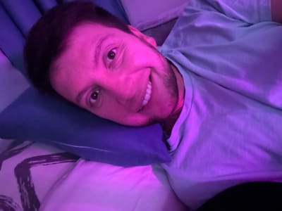 A close-up, low-angle, indoor, selfie-style shot shows a man lying on his side on a pillow, looking at the camera. He has dark hair, brown eyes, and a beard. He is smiling, showing his teeth. He is wearing a light blue t-shirt. The scene is lit with purple and pink lighting. The background shows a pillow and bedding. There is no information available about the location in the city of Tokaryovo, Russia.