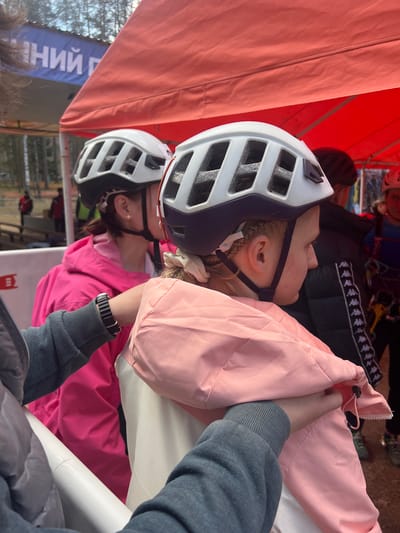 Two individuals wearing helmets are positioned beneath a tent-like structure. The location appears to be Lesnoye, Russia, based on the presence of a sign with Cyrillic text. One individual wears a pink jacket, while the other wears a pink windbreaker. A person wearing a grey sweatshirt is near the individuals, adjusting a pink jacket. A black Kappa jacket is visible in the background.