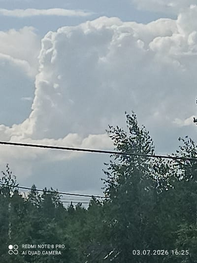 A slightly blurry, overcast sky fills the upper portion of the image, with large, white cumulus clouds dominating the frame. A dark, thin horizontal line, likely a power line, stretches across the middle of the image. Below this, a dense canopy of dark green trees forms the lower half of the scene. In the bottom left corner, white text reads "REDMI NOTE 8 PRO AI QUAD CAMERA". In the bottom right corner, white text indicates the date and time: "03.07.2026 16:25". The scene is consistent with an outdoor environment in Sovetskiy, Russia, likely during daylight hours.