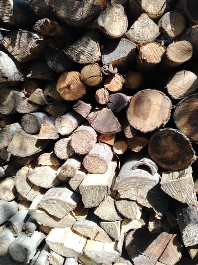 A tightly packed pile of chopped wood is shown in this outdoor, sunlit scene. The wood is of various sizes and shapes, with some pieces appearing to be split logs and others rounded sections. The texture of the wood is rough and weathered, with visible grain patterns and bark on some pieces. The lighting creates strong shadows, highlighting the contours and textures of the stacked wood. The overall impression is one of a substantial quantity of firewood, likely prepared for use. The image does not contain any people or specific identifiable landmarks within the city of Shaki, Azerbaijan, though it is a common sight in rural or suburban areas where wood is used for heating.