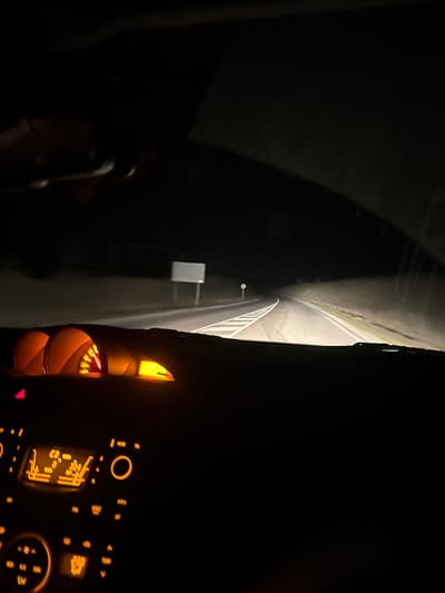 The scene depicts a nighttime view from within a vehicle, likely traversing a rural road near Malko Kirilovo, Bulgaria. The perspective is through the windshield, revealing the road ahead illuminated by the vehicle's headlights.

In the foreground, the lower portion of the vehicle's dashboard is visible. An instrument cluster, glowing with orange backlighting, is positioned on the upper left side of the dashboard, displaying gauges and a red indicator light. Below this, the central console features a climate control unit and radio interface, also illuminated with orange light, showing various buttons and digital icons.

The midground shows a two-lane asphalt road extending forward, gently curving to the right in the distance. The road surface is marked with white solid and dashed lines in the center and a solid white line on the right edge. A large, rectangular, blank white road sign mounted on two posts stands on the left side of the road, partially lit by the headlights. Further ahead on the right, a smaller circular white sign is visible. The immediate surroundings of the road are dark, appearing as low, unlit banks of earth or vegetation.

The background is entirely dark, indicating an absence of ambient light sources beyond the vehicle's illumination, consistent with a rural or undeveloped area at night. No individuals are visible within the frame, though the presence of the dashboard and illuminated controls implies an occupant is operating the vehicle. No specific actions or interactions are depicted beyond the act of driving.