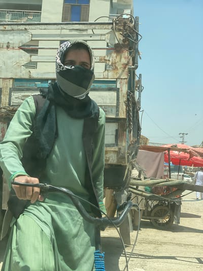 A man wearing a green tunic, a dark vest, and a scarf covering his face is on a bicycle in what appears to be a dusty urban environment, likely in or around the city of Qala-e-Naw, Afghanistan. He is positioned in front of a rusty, weathered truck. In the background, there are buildings, market stalls with red umbrellas, and other vehicles, including a cart laden with produce. The sky is clear and blue, suggesting a sunny day.