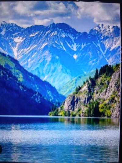 The image depicts a serene mountainous landscape with a body of water in the foreground. Snow-capped mountains dominate the background, with rugged peaks and slopes covered in patches of snow. The midground features a steep, rocky mountainside adorned with lush green vegetation and scattered trees. The foreground is a tranquil lake with rippling water, reflecting the dramatic scenery above. The sky is partly cloudy, with a mix of blue sky and white clouds. The overall impression is one of natural beauty and wilderness. No specific location in the city of Auk, Kyrgyzstan can be identified from the image.