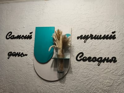 The image shows a textured wall with black Russian lettering and a decorative element. The lettering, arranged in two lines, reads "Самый день" on the left and "лучший сегодня" on the right. This translates to "The best day today". In the center, a teal-colored, rounded plaque is layered on top of a larger, light gray semi-circular plaque. A small, dark teal shelf juts out from the plaques, holding a clear glass vase filled with dried grasses and reeds, tied with twine. The wall itself has a rough, stucco-like texture. The image appears to be an interior shot. Based on the text, it's likely a decorative inscription in a cafe, restaurant, or home. There is no information to ascertain its specific location in Novyy, Russia.