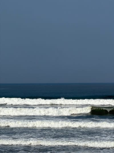 The image displays an ocean view in Belalang, Indonesia, characterized by a clear, dark blue-grey sky occupying the upper half. A distinct horizon line separates the sky from the ocean. The distant ocean water is a uniform deep blue, with a faint, singular white point visible on the far horizon, possibly a distant vessel or buoy. The lower half of the frame is dominated by multiple parallel lines of breaking waves. At least five prominent wave lines with white foam crests are visible, moving towards the viewer. The troughs between these waves show darker blue-grey water. One wave in the mid-right of the frame exhibits a pronounced curling form, revealing a darker, tubular section. No individuals or terrestrial features are visible.