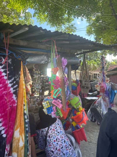 The image depicts a street-side stall in Bira, Indonesia, selling various beach-themed items and accessories. In the foreground, vibrantly colored tie-dye fabrics hang on display, a pink and black pattern on the left, followed by an orange and yellow pattern. Behind these fabrics, a variety of small toys and accessories are displayed. These include colorful plastic hair clips in shades of pink, orange, and green, as well as miniature buckets and spades, a Rubik's cube, and sunglasses. Some items are displayed in plastic packaging, while others are hung directly from the stall's structure.
The stall itself is covered by a corrugated metal roof supported by wooden beams. The background reveals more of the stall's offerings, including handbags and shell decorations, and hints at buildings and foliage beyond, typical of a coastal town. Two individuals are partially visible, one wearing a black headscarf and a patterned top, and another in a black garment on the far right. The overall scene suggests a bustling market environment with goods catering to beachgoers or tourists.