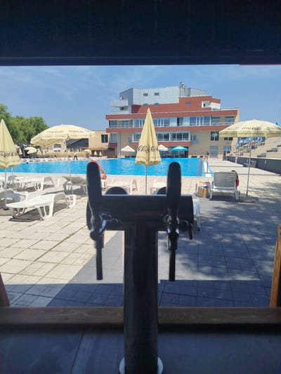 This is a medium shot of a swimming pool area, seen from behind a bar, with beer taps in the foreground. The pool is a vibrant blue, and several lounge chairs are scattered around its edge. Yellow umbrellas with "Coca-Cola" branding are positioned throughout the scene. A modern, multi-story building with a red and grey facade is visible in the background, beyond the pool. The ground is paved with light-colored tiles, and shadows suggest a sunny day. There are no people clearly visible in the image, though the context of a bar and pool area implies their presence. The scene is set in Prijedor, Bosnia and Herzegovina.