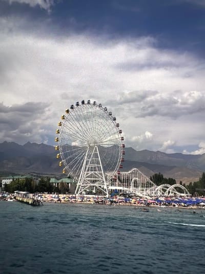 The image depicts a waterfront amusement park with a prominent Ferris wheel and roller coaster. The Ferris wheel is white with yellow and black gondolas, and the roller coaster features white tracks and supports. The park is situated along a beach with numerous beach umbrellas and people sunbathing and swimming in the water. In the background, there are mountains and buildings, suggesting an urban or semi-urban setting. The sky is cloudy, with patches of blue visible. This scene is located in the city of Kadamjay, Kyrgyzstan.