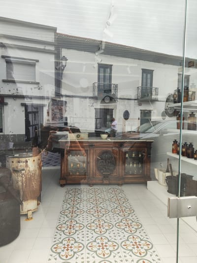 The image captures the interior of a shop in Ribeira Grande, Portugal, likely an apothecary or antique store, seen through a glass storefront. The interior features decorative tiled flooring, a large wooden cabinet with glass display cases, and shelves stocked with antique apothecary bottles and equipment. In the reflection of the glass storefront, a section of the street is visible, showing buildings with balconies, including architectural details characteristic of Ribeira Grande, and a person standing by a car. Another person is reflected holding a camera in front of the store, capturing the shop's display.