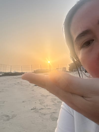 A close-up selfie captures a person's face and hand on a beach in Ajman, United Arab Emirates, during sunset. The sun is low on the horizon, casting a warm, orange glow across the sky and sand. The person's left hand is held up in the foreground, obscuring part of the view of the beach and the setting sun. The beach appears to be sandy with some footprints visible. In the background, there are fences and structures, possibly part of a construction site or resort. The overall scene evokes a sense of a peaceful evening by the sea.