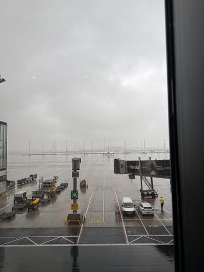 The image depicts an airport apron, most likely at Berlin Brandenburg Airport (BER) in Schönefeld, Germany, observed through a window. The weather is overcast and wet. Visible are parked vehicles including vans and baggage carriers. A person wearing a high-visibility jacket is standing on the apron near the jet bridge branded with "Revolut" advertisement. Aircraft and runway markings are visible in the background. The gate number is indicated as B14.