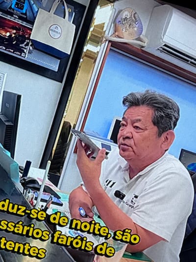 The image captures a man of East Asian descent with graying hair, wearing a white polo shirt with a logo that includes the text "to Girage". He is holding a silver object, possibly a hair clipper, in his right hand and appears to be talking. In the background, a white air conditioning unit is visible mounted high on a wall. Below the man, text in Portuguese is partially visible: "duz-se de noit", "ssários faróis de", and "tentes". On the left side of the image, a framed picture displays a tote bag with a circular logo. The overall setting suggests an indoor environment, possibly a salon or a workshop. The location in the city of Almada, Portugal is not evident from the image.