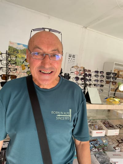 A middle-aged Caucasian man with a shaved head is standing in an optical shop, possibly in Cherkasy, Ukraine, as indicated by the Cyrillic text on the eye chart and the name "FEDOROV" on a box. He is wearing glasses on his face and another pair on his head. He has a teal-colored t-shirt with the text "BORN IN ALPS SINCE 1973" printed on it and a black strap across his chest. The background features shelves stocked with various eyeglasses, sunglasses, and eye care products. A calendar for 2023 hangs on the wall, displaying a landscape image. The lighting suggests an indoor setting, likely during the day.