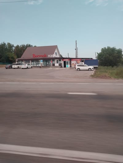 The image depicts a scene along a road in Баюновские Ключи, Russia. A building with a reddish-brown pitched roof, identified by the Russian text "Восток кафе" (Vostok Cafe) in red lettering, is visible on the left side of the road. The building appears to be a cafe or restaurant, with a large glass storefront. To the right of the cafe, several cars are parked. A white van is parked in front of the cafe, and two white cars are parked in a line behind it. A dark-colored car is also partially visible behind the white cars.
Behind the cafe, a dark blue truck is parked. The area around the cafe is paved, suggesting a parking lot or roadside rest stop. A grassy area with trees borders the road and the cafe. The road itself has white lane markings and appears to be a paved highway. The sky is light blue with some wispy clouds, indicating it is daytime, likely in the late afternoon or early evening given the soft lighting. The overall impression is of a casual roadside establishment, possibly a place for travelers to stop and eat. No people are discernible in the image. The image has been rotated 90 degrees clockwise, making the text appear vertically oriented.