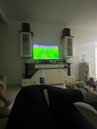 An indoor scene, likely a living room, in Episkopi, Cyprus, is depicted. The central focus is a large flat-screen television displaying a soccer match on a green field with numerous players. A digital scoreboard in the upper left corner indicates "1:18," a score of "0-0," and "2nd HALF." The television is mounted above a light-colored entertainment unit with dark accents. Two wall-mounted cabinets, each topped with a dark speaker, flank the television. To the far right, a water cooler with a large clear water bottle is visible.

In the immediate foreground, the lower body of an individual is visible, seated or reclined, wearing dark trousers and light-colored socks or footwear on the right foot. A white-handled knife with a metallic blade is resting horizontally on the person's upper thigh or lap, with the blade pointing towards the right. On a clear glass coffee table in front of the individual, a blue-labeled Pepsi bottle stands upright. Multiple light-colored bowls, a clear drinking glass, and several metallic beverage cans (red and silver) are also present on the table.

To the far left, a small, light-colored dog is partially visible, positioned next to a green armchair and facing towards the television. The room features light-colored walls and a light-colored tiled floor, and is moderately lit.
