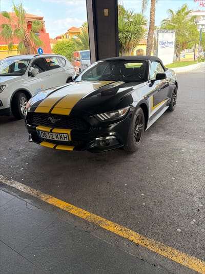 A black convertible car with yellow stripes is parked in what appears to be a parking area. A white car with the logo of "Islas Canarias" is parked beside it. The license plate of the convertible is "0612 KKH." Outside the parking area, palm trees are visible, along with buildings and a sign that reads "Best Tenerife." The scene likely takes place in Los Cristianos, Spain, given the presence of the Best Tenerife hotel, which has a branch in that city. A yellow line is painted on the road adjacent to the parking space. The image appears to be taken from inside a building looking outwards.