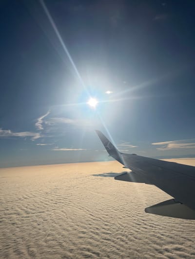 An aerial image, presumably taken over Ilshofen, Germany, captures a view from an aircraft window showing the right wing of the airplane in the foreground, extending from the middle-right to the bottom-right of the frame. The winglet prominently displays the Lufthansa crane logo. Below the aircraft, an extensive, continuous layer of textured, white and light beige clouds, resembling stratocumulus, dominates the lower half of the scene, stretching to a distant, hazy horizon. The upper half of the image features a clear, deep blue sky, with the sun positioned in the upper-center to upper-left quadrant. The sun emits a strong starburst lens flare and a bright halo, indicating direct illumination. A thin, wispy cloud formation is visible in the sky to the left of the sun. No people, specific objects on the ground, or discernible interactions are visible due to the high altitude and complete cloud cover.