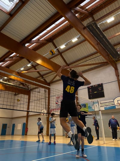 The image depicts an indoor volleyball practice or game in a gymnasium in Sennecey-le-Grand, France.

In the foreground, a male player is captured mid-air, having jumped to spike a yellow and blue volleyball positioned above him. He faces away from the camera, wearing a dark blue t-shirt with "SUC Volley Ral" and the yellow number "48" printed on the back, black shorts, and athletic shoes with prominent white and black soles. His arms are raised above his head, hands open in preparation to strike the ball. Directly below him, another male player in a dark blue t-shirt with the number "8" stands on the blue-marked court, looking upwards.

Across the black volleyball net, on the left side of the frame, two young male players are visible. One stands slightly bent, while the other stands upright, both wearing light blue and white jerseys with numbers and shorts. A second yellow and blue volleyball rests on their side of the court near the net.

In the background on the right, two adult males observe the scene. One, dressed in a dark tracksuit with red trim, stands near a wall-mounted digital scoreboard displaying "LOC" and "VISIT" above numeric values. The other, wearing a dark blue polo shirt with white trim and dark pants, holds a yellow and blue volleyball.

The gymnasium features a light-colored wooden floor marked with blue and white lines indicating court boundaries. The high ceiling is characterized by exposed, stained wooden beams supporting a white metal roof deck, with multiple elongated fluorescent light fixtures and visible skylights. Various French-language banners and posters are affixed to the white walls, one partially visible banner reads: "Communauté de Communes Entre Saône et Grosne soutient le Sport & la Culture." Another partially visible sign reads "TOUS TRAVAUX J[...]" with "Gouleur" below it.