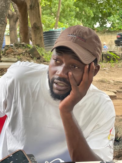 The image shows a close-up, portrait-style shot of a Black man wearing a brown baseball cap and a white t-shirt. He is resting his head on his hand, with his fingers splayed across his forehead and temple, obscuring part of his face. His eyes are open and looking towards the camera. He has a beard and a visible smile, with his teeth showing.
The background appears to be outdoors, with lush green foliage on the left side of the frame. There are also some wooden structures and what looks like a large black water tank or barrel behind him, suggesting an informal or rural setting. The lighting suggests it could be daytime, possibly overcast or in shade.
The man's t-shirt has some text and graphics on it, but they are partially obscured and difficult to read clearly. The cap has a logo embroidered on it, which appears to be red, white, and blue stripes with text above them. The overall mood of the image is relaxed and candid.
The scene is set in Wupa, Nigeria, suggesting a local context.