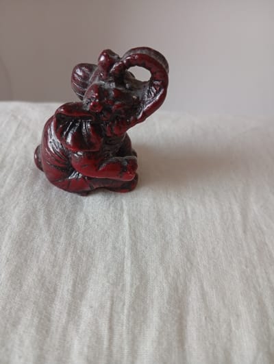 A dark red, carved figurine of an elephant sits on a textured white cloth. The elephant is small and appears to be made of wood or resin. Its trunk is raised and curved, and it is depicted in a seated position. The background is a plain, light-colored wall. There is no indication of the location being in Civitanova Marche, Italy.