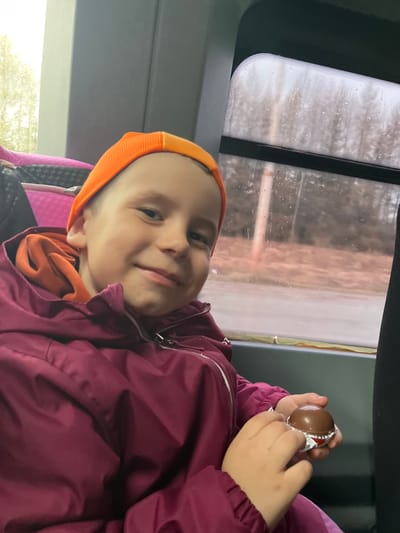 A child in a car, possibly in Novoe Mart'yanovo, Russia, is holding a chocolate egg wrapped in foil. The child is wearing an orange hat and a burgundy jacket. The background shows a car window with a blurred outdoor scene.