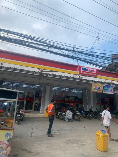 The image shows the exterior of an Alfamart convenience store located in Lagadar, Indonesia (latitude: -6.921, longitude: 107.539). The store has a red and yellow awning and large glass windows. Numerous electrical wires are visible above the store. There are several motorcycles parked in front of the store, along with a food stand and a yellow trash can. Four people are present in the image, including a man in an orange vest standing to the left of the frame. Another man in a white shirt is walking to the right. Two additional men are near the motorcycles, appearing to work on one of the motorcycles. The pavement surrounding the store is cracked. A nearby building is visible in the background. The sky is cloudy with a light blue hue.