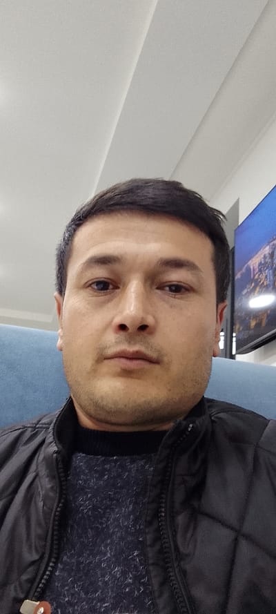 The image presents a headshot-style portrait of an adult male in an indoor setting within the city of Chinabad, Uzbekistan. The subject, facing forward with a neutral expression and direct gaze, occupies the majority of the frame. He has short, dark hair, brown eyes, a straight nose, full lips, and light stubble on his chin and jawline. He is dressed in a dark, possibly black, quilted jacket unzipped over a dark blue, textured knitted sweater. Behind him, a light blue upholstered surface, likely a chair or sofa, is visible to his left. The upper background features a light-colored ceiling with a diagonal seam. To the subject's right, a portion of a black-framed television screen displays an illuminated night cityscape.