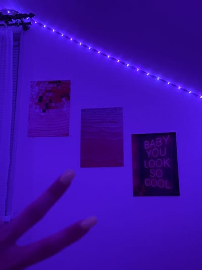 The image shows a wall decorated with three framed pieces of art. From left to right, the first piece is a rectangular artwork with a mosaic pattern of squares in shades of pink, red, and black. The second piece is a rectangular artwork depicting a body of water with subtle wave patterns, primarily in shades of pink. The third artwork is a neon sign that reads "BABY YOU LOOK SO COOL" in pink lettering against a dark background. The wall is illuminated by purple LED string lights that run along the top edge. A hand with painted nails is visible in the foreground, with two fingers extended upwards, forming a peace sign. The scene appears to be indoors, likely a bedroom or personal space, and does not contain any discernible elements that would associate it with the city of Obninsk, Russia.