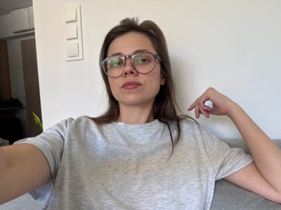 The image shows a woman wearing glasses and a grey t-shirt. Her head is tilted to the side, and her hair is dark brown and falls around her face. The woman is looking at the camera, and her mouth is slightly open. The background is a plain white wall with a light switch. The image appears to be taken indoors, possibly in a home or apartment. The lighting suggests it is daytime. There is no visible text in the image. The overall impression is a casual, candid portrait.
