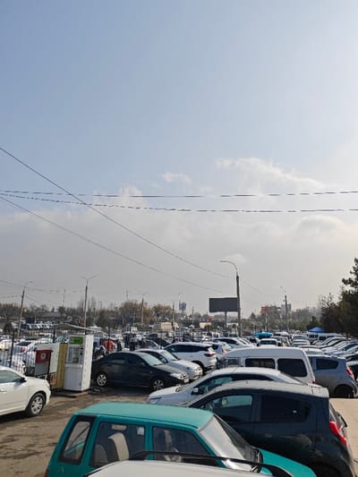 This image depicts a densely packed outdoor parking lot in Eshanguzar, Uzbekistan, under a light blue sky with scattered white clouds. Multiple dark utility lines crisscross the upper and middle portions of the frame. The paved surface, likely asphalt, is filled with numerous automobiles of various makes and models, including sedans, hatchbacks, SUVs, and a white van, parked in rows from the immediate foreground to the mid-ground. Prominent vehicles include a teal/turquoise compact car in the immediate foreground, a white sedan to the left, and a dark grey hatchback on the right. In the mid-ground, two tall, light-colored kiosks or dispensers are visible on the left side of the parking area. Multiple streetlights or utility poles are scattered throughout the lot and background. A large, dark, rectangular billboard structure is present in the mid-background. Indistinct human figures are observed walking or standing among the vehicles in the mid-ground. The background features a line of deciduous trees, some appearing bare, suggesting late fall or winter, along with various buildings and an elevated structure, possibly a bridge, in the distance. The scene is illuminated by natural daylight.