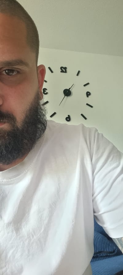 The image shows a close-up of a man's face and torso, with a wall clock in the background. The man has a short haircut and a full beard. He is wearing a white t-shirt, which has some faint stains on the front. The wall clock is a modern, minimalist design with black numbers and hands on a white background. The time on the clock is approximately 10:55. The background appears to be an interior wall, painted a light color. The overall lighting suggests an indoor environment, possibly a home. The location is specified as Mehltheuer, Germany.