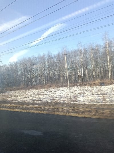 The image depicts an outdoor winter or early spring landscape in Drezna, Russia. The scene is viewed under a light blue sky with faint, linear white contrails or cirrus clouds. Multiple dark, parallel utility lines span horizontally across the upper portion of the frame, with a large, dark utility tower partially visible on the far left. The background consists of a dense, leafless forest composed primarily of slender deciduous trees, many with the pale trunks characteristic of birch. In the midground, an open field shows an uneven distribution of melting snow patches interspersed with exposed brown earth and dry vegetation. A single, tall, light-colored utility pole stands vertically, slightly leaning, in the right midground. The foreground is a dark, rough surface, possibly a road shoulder or unpaved ground, exhibiting a small puddle on the left and a faint, horizontal tan line across its width. No individuals or active subjects are present within the frame.