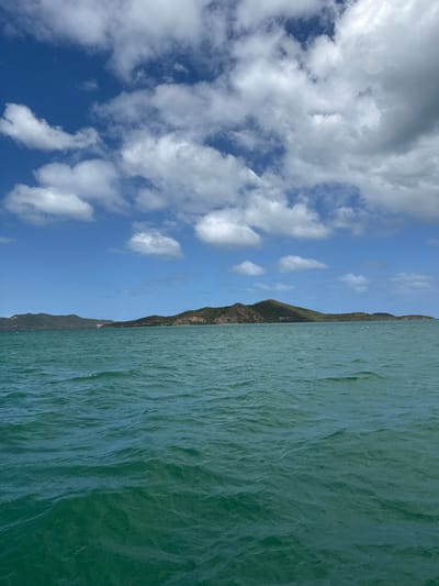 The image displays an expansive outdoor seascape in Dumbéa, France, under a clear blue sky dotted with numerous white cumulus clouds. The lower two-thirds of the frame are occupied by a body of teal-colored water, its surface characterized by consistent small ripples and waves. Stretching horizontally across the middle ground is a distant, vegetated landmass, appearing as an island or coastline. This landmass exhibits varied topography, featuring multiple green, tree-covered hills and visible areas of exposed rock, particularly on the prominent central peak. No individuals, vessels, or artificial structures are discernible within the frame. The scene is illuminated by bright, natural daylight.