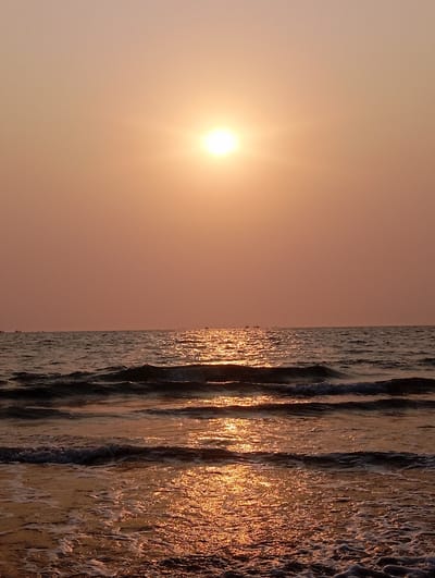A serene seascape is captured at either sunrise or sunset, dominated by a warm, golden light. The sun, a brilliant white orb with visible rays, is positioned centrally in the upper part of the frame, illuminating the scene. The sky above is a gradient of soft orange and reddish-pink, fading slightly towards the top.

The vast expanse of the ocean fills the lower two-thirds of the image. A shimmering path of golden light, created by the sun's reflection, stretches across the water towards the viewer. This intense reflection highlights the crests of the gentle waves, making them appear to glow with a molten gold hue. Further out, the ocean appears a darker blue-grey where it is not directly illuminated.

Several waves are visible, moving towards what appears to be a shore just beyond the bottom of the frame. White foam marks the breaking edges of the closer waves, indicating their gentle movement. On the distant horizon line, faint silhouettes of a few small vessels, likely fishing boats, can be discerned against the bright sky and water. There are no people visible in the image. The overall impression is one of tranquility and natural beauty, characteristic of a coastal setting like Arambol, India, during the golden hour. No text is visible in the image.
