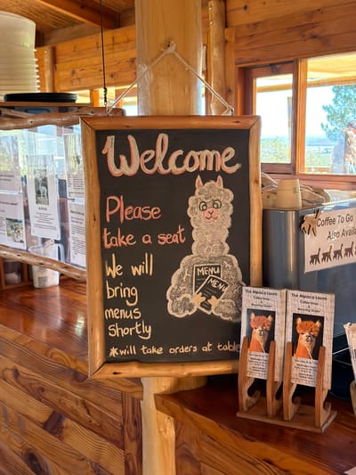 This media file captures the inviting interior of a rustic establishment, likely a coffee shop or weaving studio, characterized by extensive use of natural wood for walls, furniture, and window frames.

The central subject is a black chalkboard sign, edged with natural wood, prominently displaying a "Welcome" message in red and white chalk. Below, the sign instructs: "Please take a seat We will bring menus Shortly *will take orders at table." A charming, fluffy alpaca character, drawn in chalk, holds a "MENU" sign, reinforcing the business's theme.

To the right of the chalkboard, a wooden display stand holds several brochures titled "The Alpaca Loom Coffee Shop & Weaving Studio." These brochures advertise "Open Daily 09:30 - 17:30," "Alpaca Barn Tours Every hour on the hour," and "Alpaca Walks: 10:00 & 16:00." Further right and slightly behind, another sign indicates "Coffee To Go Also Available" and features silhouettes of alpacas.

In the background, large wooden-framed windows reveal bright daylight and glimpses of green trees, suggesting a clear, sunny day. The overall setting is warm and natural, implying a tranquil, welcoming atmosphere. The visible text clearly indicates that the establishment is "The Alpaca Loom," offering coffee, a weaving studio, and alpaca-related experiences. This fits the location context of Drakenstein Nu, South Africa, suggesting a farm or nature-based visitor attraction.