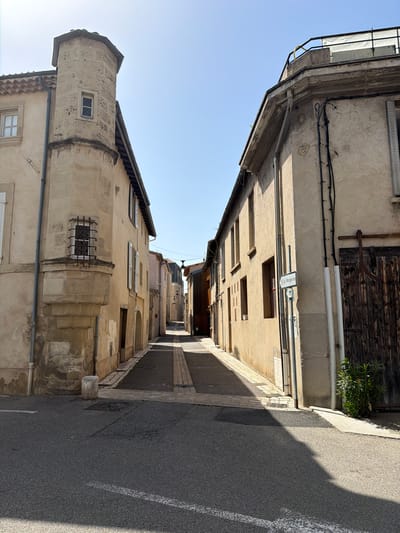 This is a vertical, medium-shot, outdoor photograph captured on a clear, sunny day in Mercurol-Veaunes, France. The scene depicts a narrow, paved street winding between two aged, light-colored buildings under a bright blue sky.

On the left, the building features a distinct, rounded stone tower with a small, barred window and a tiled roof. Adjacent to the tower, the building has several windows with white shutters and a grey downspout. The wall exhibits signs of age and discoloration.

On the right, the building is also constructed from a light-colored material, with visible electrical conduits running vertically along its facade. It has a few rectangular windows and a large, weathered wooden door with metal detailing. A small sign with green lettering and an arrow points to "Le Mangevins". A thin, green plant grows near the base of this building.

The street itself is paved with asphalt, marked with a white line, and lined with cobblestones. Sunlight casts distinct shadows across the street and the buildings. In the distance, the street continues, lined with more buildings and street lamps. There are no people visible in the image. The overall impression is one of a quiet, historic street in a French town.
