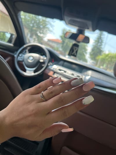 The image displays a person's right hand, adorned with a manicure and rings, positioned inside a car with a bright outdoor background.

**Subjects:** The primary subject is a human right hand. Elements of a car interior and blurred exterior scenery are secondary subjects.

**Objects:** The hand features square-shaped nails painted in a varied style: pale pink on the pinky and middle fingers, sparkling silver glitter on the ring finger, and off-white with a line of small rhinestones near the cuticle on the index finger. On the ring finger, a prominent ring with a central stone flanked by smaller stones is worn, accompanied by a thinner band underneath it. The car's steering wheel, clearly displaying the BMW logo, is in the blurred mid-ground. Parts of the dashboard, a rear-view mirror, and a side mirror are also visible.

**Scene/Setting:** The scene is set inside a modern car, identified as a BMW. The car appears to be stationary or moving slowly, allowing for a clear focus on the hand. Through the car windows, a bright, sunny outdoor environment with blurred green trees and foliage is visible. The provided location context is Madara, Bulgaria.

**Activity or event taking place:** The image captures the casual display of a recent manicure and a ring, likely a personal snapshot taken while traveling or waiting in the car.

**Notable details:** The photo is taken in bright daylight, indicating a sunny day. The manicure exhibits a mix of subtle and glittery nail polish, with decorative rhinestones. The focus is sharp on the hand, while the background is intentionally blurred to highlight the subject.

**Visible text:** The "BMW" logo is clearly identifiable on the steering wheel. A faint, blurry white "X" on a dark background is discernible in the reflection within the rear-view mirror.