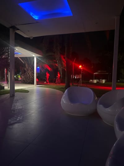 The scene depicts an outdoor night event, likely in Santa María, Argentina, though the precise location within the city is not discernible from the image. The perspective is from under a modern white overhead structure, featuring a recessed rectangular ceiling panel emitting blue light. The immediate foreground shows a smooth, light-colored paved surface, upon which at least three white, spherical lounge chairs are situated, one prominently in the mid-right. Beyond the paved area, a grassy expanse is visible, bordered by multiple palm trees. Approximately five to seven individuals are present on this grassy area. Two figures are located in the left-midground, illuminated by red light, appearing to walk or stand. To their right, a group of three to five individuals stands near a tripod-mounted light fixture that emits a red light or flash. Further in the background, a white canopy or tent structure is visible, surrounded by several low-profile white seating objects. The environment is predominantly dark, illuminated by artificial blue and red accent lighting, creating strong contrasts and shadows.