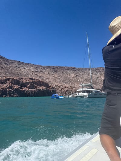 A view from a boat on the Sea of Cortez in Baja California, Mexico. The sea is turquoise with white water from the boat's movement. A rocky, arid cliff is to the left. Several sailboats are anchored at the base of the cliff, including one catamaran named "CARPE DIEM". A portion of a person wearing a straw hat, black shirt, and gray shorts is in the top portion of the frame. The sky is blue.