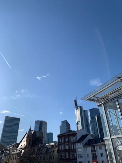 The image shows a view of the Frankfurt skyline from the ground level. The image is taken in the city of Frankfurt am Main, Germany at latitude 50.108 and longitude 8.685. The view is from the street level looking up at a cluster of skyscrapers. The view is partially obstructed by a glass and metal canopy. On the right side of the image, two modern skyscrapers are visible. On the left side of the image, the tops of several modern skyscrapers are visible. In the foreground, the roofs of several older buildings are visible. The sky is blue with some white clouds. There is a contrail in the sky. The image is taken on a sunny day. There is a streetlight on the left side of the image. There are no people in the image. There are a few trees and plants visible. The image is taken from a low angle.