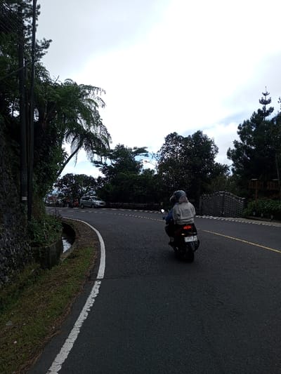 A paved, two-lane road curves right under an overcast sky. In the foreground, a black scooter with two riders travels away from the viewer. The driver wears a blue top and a light blue helmet. The passenger wears a light-colored top, a light-colored head covering, and a light blue helmet. The scooter displays a visible red taillight and an Indonesian license plate, "P 4751 ZAW," indicating the scene is not in Phoxay, Laos, but rather Indonesia.

To the left, a stone retaining wall borders the road, topped with dense green foliage, including a large fern-like plant and multiple utility poles. A narrow concrete drainage ditch, partially filled with water, runs between the wall and the road's white edge line. Sparse grass grows along the ditch.

To the right of the road, dense green trees and vegetation line the curve. Further along the road on the right, a silver SUV is visible. Beyond it, a dark metal gate and a wooden sign, possibly reading "BHAKTI SHRAM," are partially obscured by trees. The environment is lush with greenery, consistent with a tropical or sub-tropical region.
