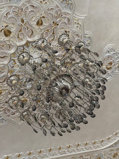 A close-up, eye-level shot captures a crystal chandelier hanging from a highly ornate, plaster ceiling. The ceiling features intricate relief patterns with delicate, flowing curves and floral motifs, accented with gold leaf. The chandelier itself is made of polished silver-toned metal and adorned with numerous faceted crystals of various sizes, some clear and some appearing to have a smoky gray tint. The crystals hang in cascading strands from the chandelier's metallic arms and decorative elements. The lighting in the image is soft, reflecting off the metallic surfaces and crystals. The scene is indoors, likely in a room with elaborate decorative architecture, such as a palace, mosque, or high-end establishment in Urgench, Uzbekistan, given the stylistic resemblance to historical Islamic architectural ornamentation. There are no people visible in the image.