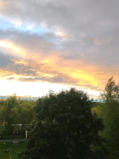 This is a medium shot, eye-level photograph of a park-like setting with a dramatic sky. The main subjects are the lush green trees in the foreground and midground, with a prominent, large, dark green tree dominating the center and right of the frame. Behind the trees, the sky is filled with colorful clouds illuminated by the setting sun, casting a warm, golden-orange glow across the scene. The sky transitions from these warm hues to cooler blues and grays at the top. Streaks of rain are visible, indicating a light shower.

In the background, a fence surrounds a grassy area with some playground equipment, including what appears to be a small slide or play structure with red and gray components, and yellow benches or seats. A yellow lawnmower or similar equipment is also visible on the grass. The presence of a fenced-in play area and well-maintained trees suggests a residential park or backyard in a suburban or urban environment.

The time of day is clearly sunset, given the vibrant colors in the sky. The weather is a mix of rain and sunshine, creating a potentially moody or picturesque atmosphere. The overall emotion conveyed is one of natural beauty and perhaps tranquility, despite the presence of rain. There are no visible text elements in the image. The location is identified as Сокол, Russia.