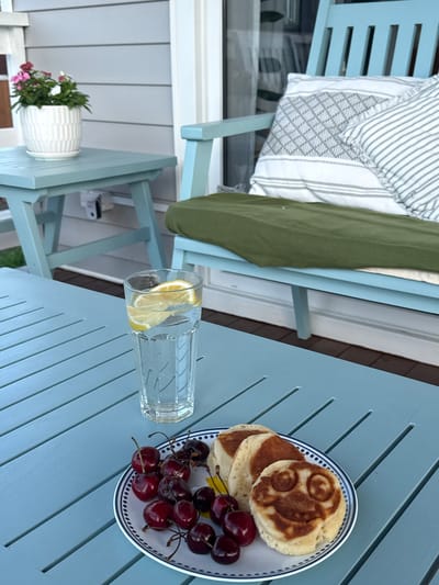 A close-up, top-down shot shows a light blue slatted outdoor table with a meal. On the right side of the table is a white plate with a blue and white striped rim. On the plate are several small, round pancakes, some with slightly darker, swirled markings on top, and a pile of dark red cherries. To the left of the plate is a clear glass filled with water and two slices of lemon. In the background, to the left, is a light blue chair with a striped cushion and a green throw blanket. The scene appears to be outdoors, possibly on a patio or deck, given the table and chair. The lighting suggests it is daytime, possibly morning or afternoon. There is no visible text in the image.