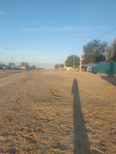 A long, unpaved road stretches into the distance, lined with houses and trees on both sides. The road surface is sandy and appears to be a dirt track with tire marks. In the foreground, a long shadow of a person is cast across the road, indicating they are standing in the foreground facing away from the camera. The sky is blue with a few wispy clouds and the moon is visible in the upper left quadrant. The scene depicts a rural or semi-rural environment, consistent with the outskirts of a city like Aral, Kazakhstan, known for its desert climate and unpaved roads. The lighting suggests it is either early morning or late afternoon.
