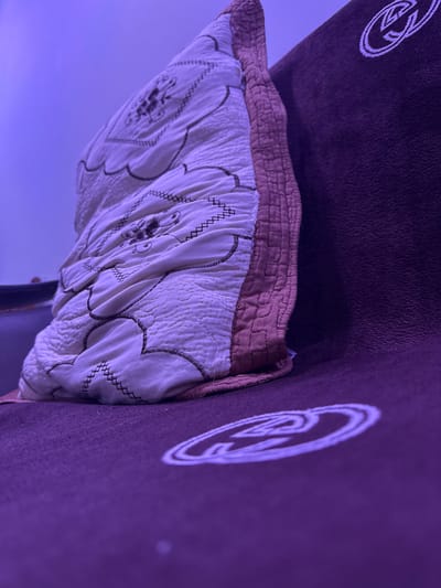 The image shows a close-up of a pillow and bedding, possibly on a bed or sofa. The pillow is a light color, possibly white or cream, and features quilted stitching creating a textured surface. Darker thread is used to outline floral or organic shapes and to create a zigzag stitch pattern along the edges of these shapes. The pillow also has a band of brown fabric along its bottom edge, which is also quilted with a cross-hatch pattern. This is layered on top of a darker, textured fabric, likely the main bedding or a throw. A prominent circular logo with interwoven lines, appearing to be stylized letters or a symbol, is visible in white on the dark fabric to the left and right sides of the frame. The scene is illuminated by a purple light source, casting a distinct hue over the entire image and creating a moody or atmospheric effect. The background is a soft, blurred gradient of purple and blue, suggesting a dimly lit room or a stylized environment. There is no discernible time of day or weather information, nor any specific location cues beyond the provided Mastaga, Azerbaijan context. The image focuses on the textures and patterns of the bedding and the illuminated logo, giving it an abstract or artistic feel. No people are visible, and no specific activity is taking place.